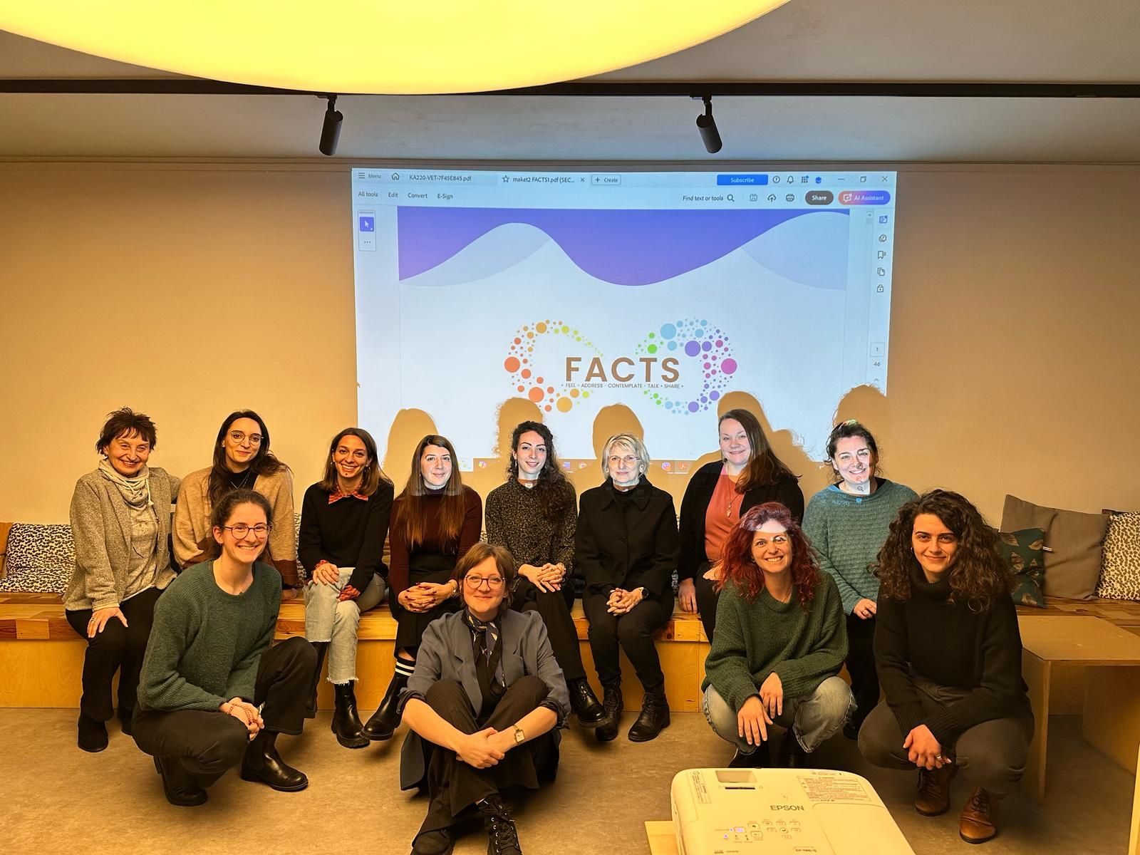 A group of people sitting on the floor in front of a screen showing the logo of the FACTS project.